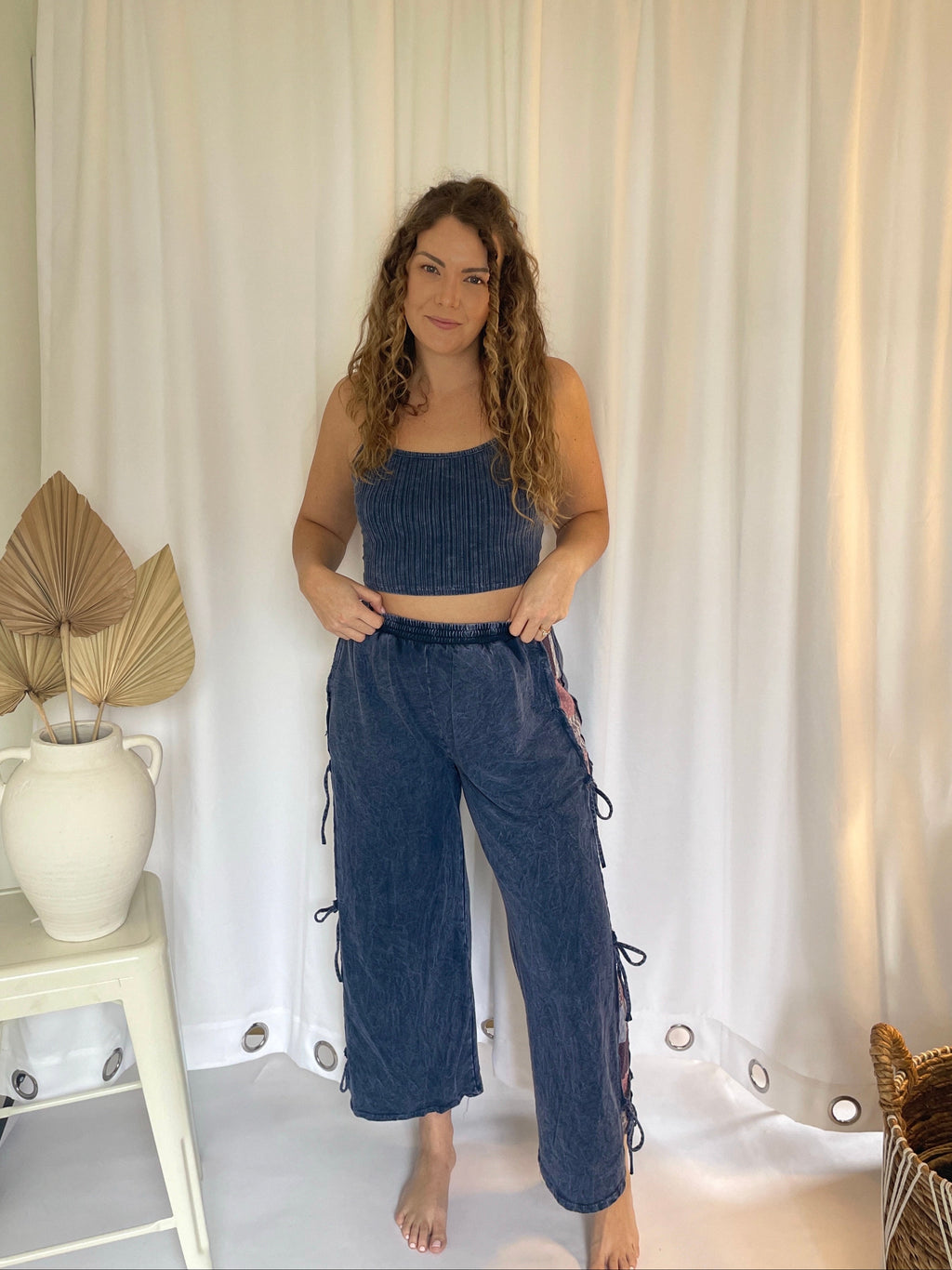 Woman wearing a blue outfit standing in front of white curtains with a plant and small table in the background.