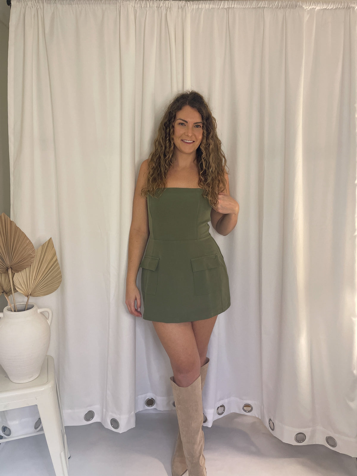 Woman in a green dress standing in front of white curtains with a plant on a table to her left.