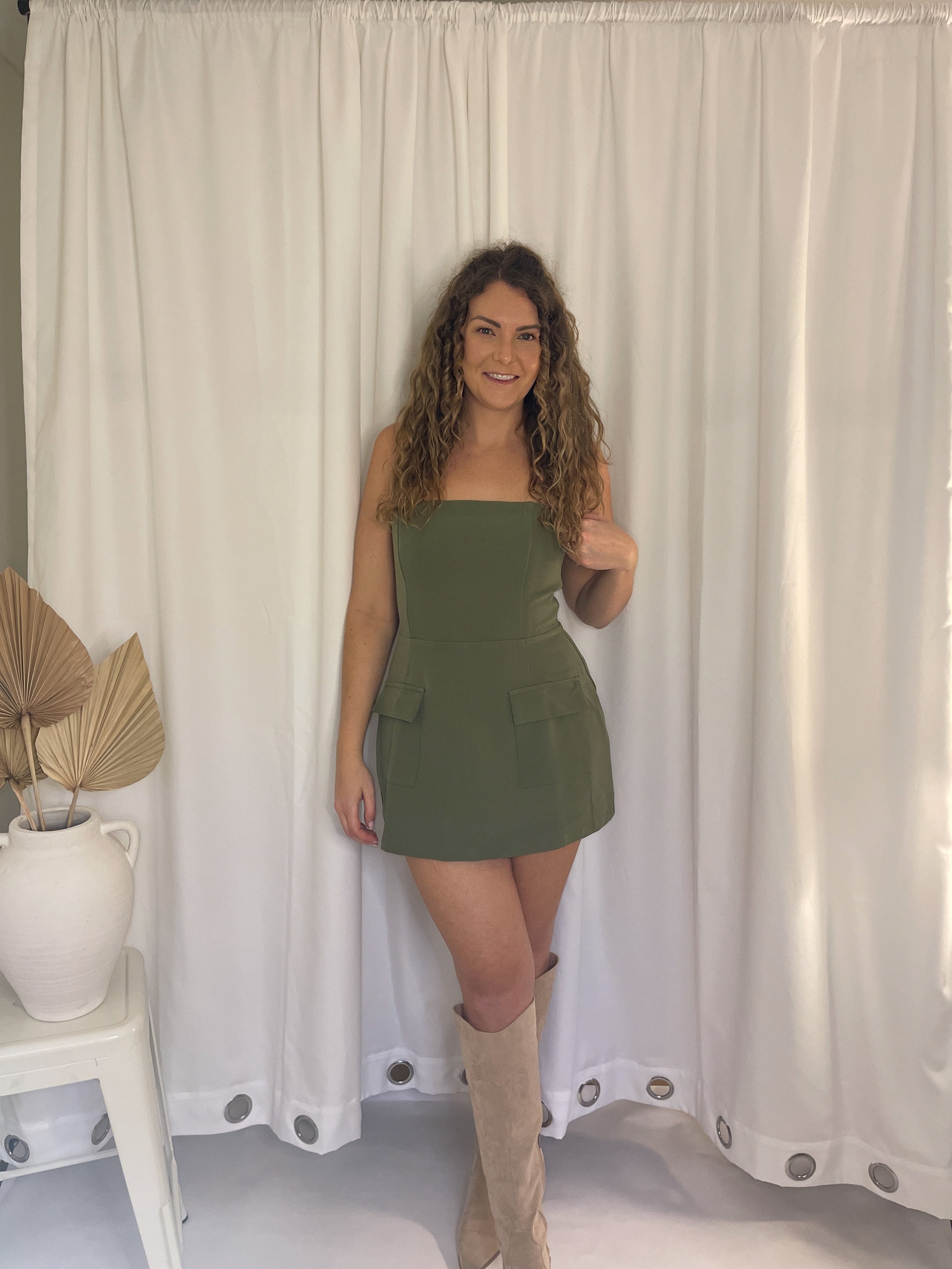 Woman in a green dress standing in front of white curtains with a plant on a table to her left.