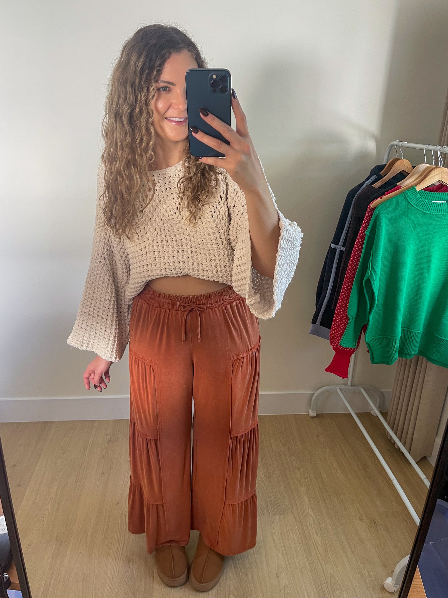 Woman taking a mirror selfie wearing a beige top and rust-colored pants in a room with clothing racks.